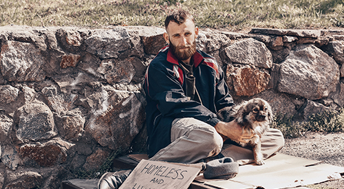 A homeless man with his dog next to a stone wall